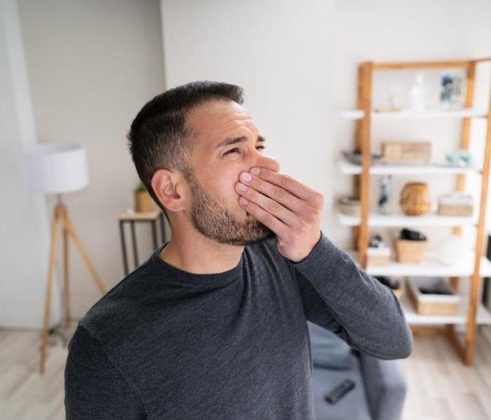 A man standing in a living room, holding his nose to avoid a bad smell.
