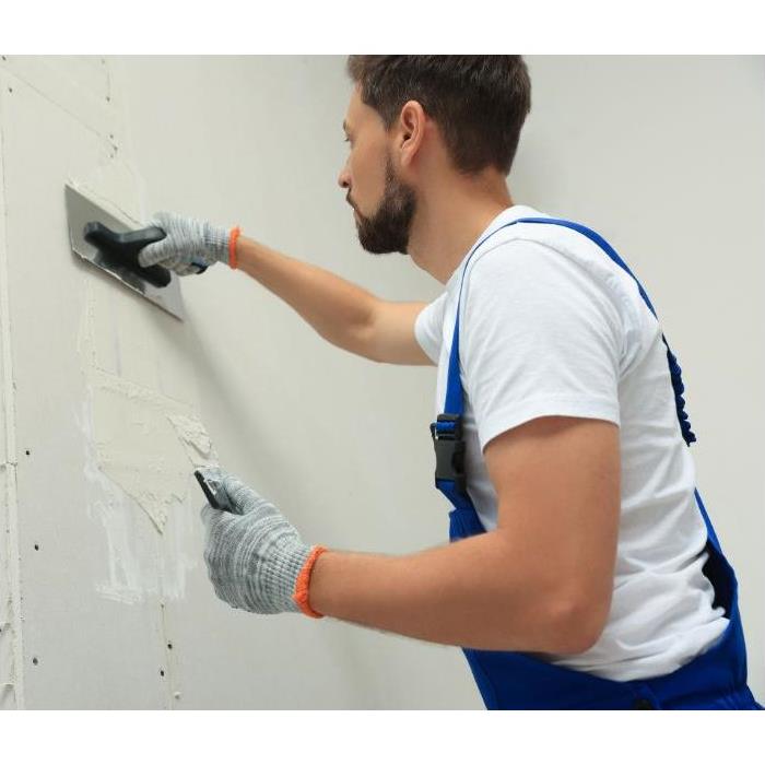 A man wearing blue overalls smooths drywall mud on a wall.
